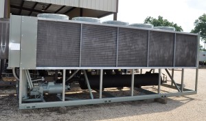 Large industrial chiller unit with multiple fans on top, situated outdoors on gravel near a metal building.