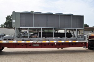 Large industrial HVAC unit on a flatbed trailer, featuring industrial chiller parts, positioned outdoors near a building and trees.