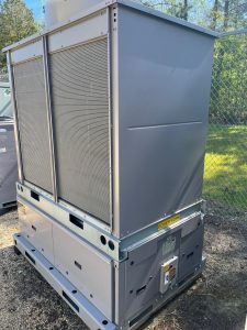 A large industrial chiller stands outdoors on gravel, surrounded by a metal fence and trees, with warning labels visible on its side.